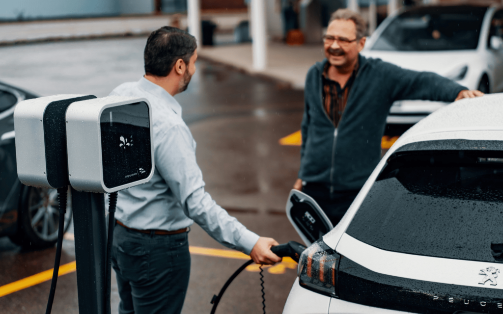 Two men are standing near an electric vehicle, with one of them connecting the charging cable to the car at an outdoor charging station.