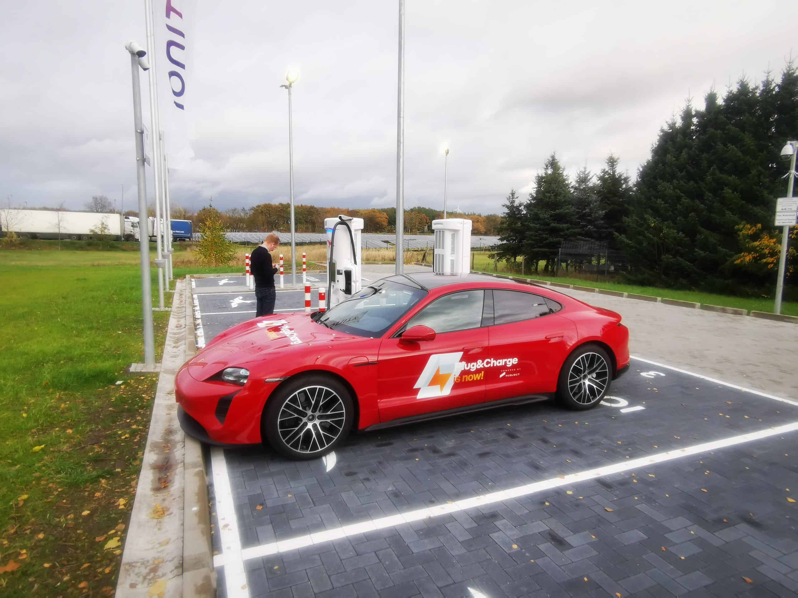 A red sports car is parked at an electric vehicle charging station. The car has a logo featuring a lightning bolt and the text "Plug&Charge." A person is seen in the background near the charging unit. The scene is set in an open area with green grass and trees.