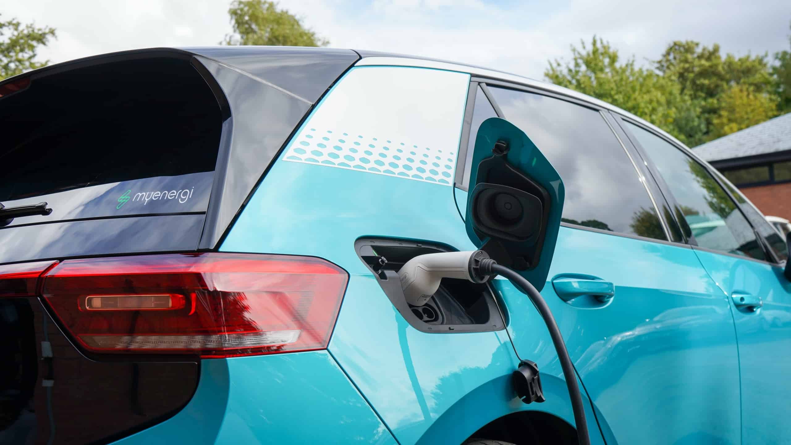 A turquoise electric car is being charged at a charging station. The rear left side of the car is visible, with the charging port open and a charging cable plugged in. Trees and part of a building are seen in the background.